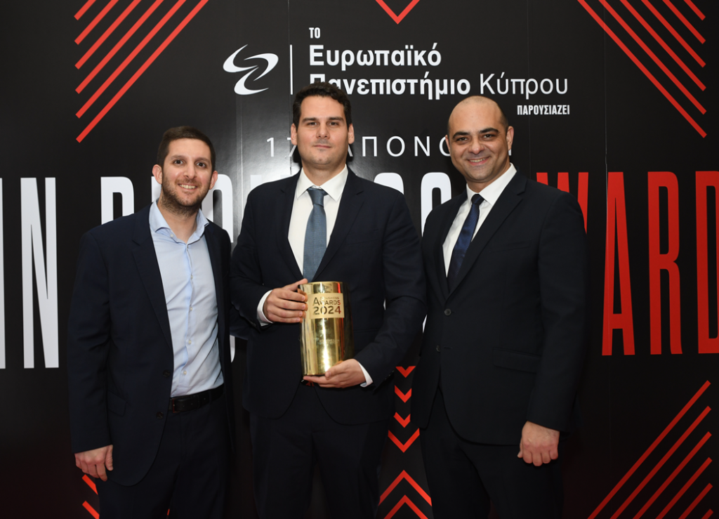 Andreas Vassilopoulos stands next to Michael Tyrimos, CEO of Capacitor Partners, who is holding a gold InBusiness Awards 2024 trophy, flanked by another executive, in front of the event’s branded backdrop.
